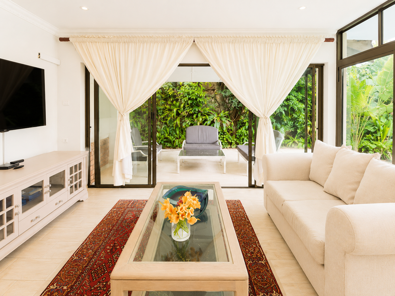 Bright living room featuring large sliding glass doors that open onto a lush garden patio, complemented by neutral tiled flooring and recessed lighting.