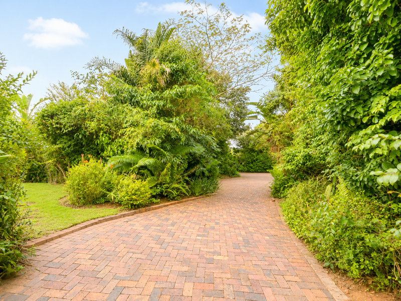 A well-maintained brick paved driveway winding through a lush, mature landscaped garden with established trees and greenery.