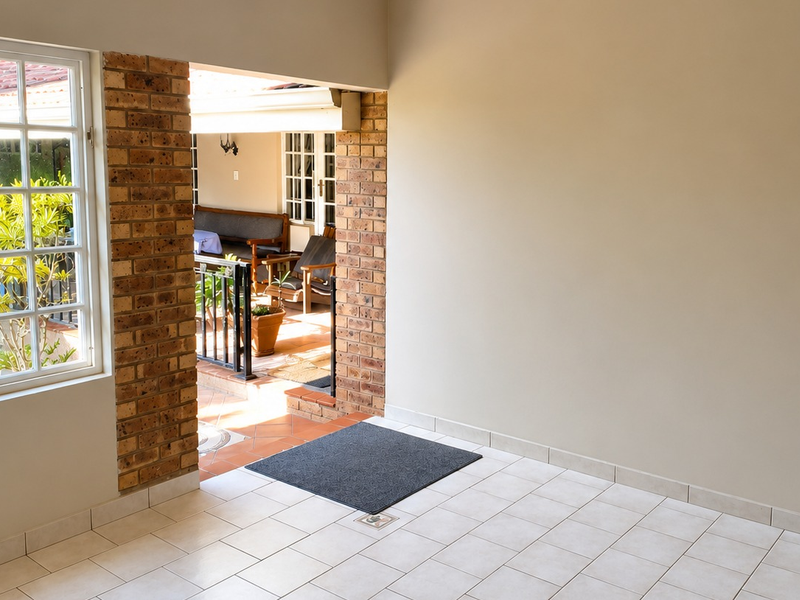 A clean, covered patio area featuring exposed brick pillars, tiled flooring, and a large multi-pane window looking out toward an adjacent outdoor space.