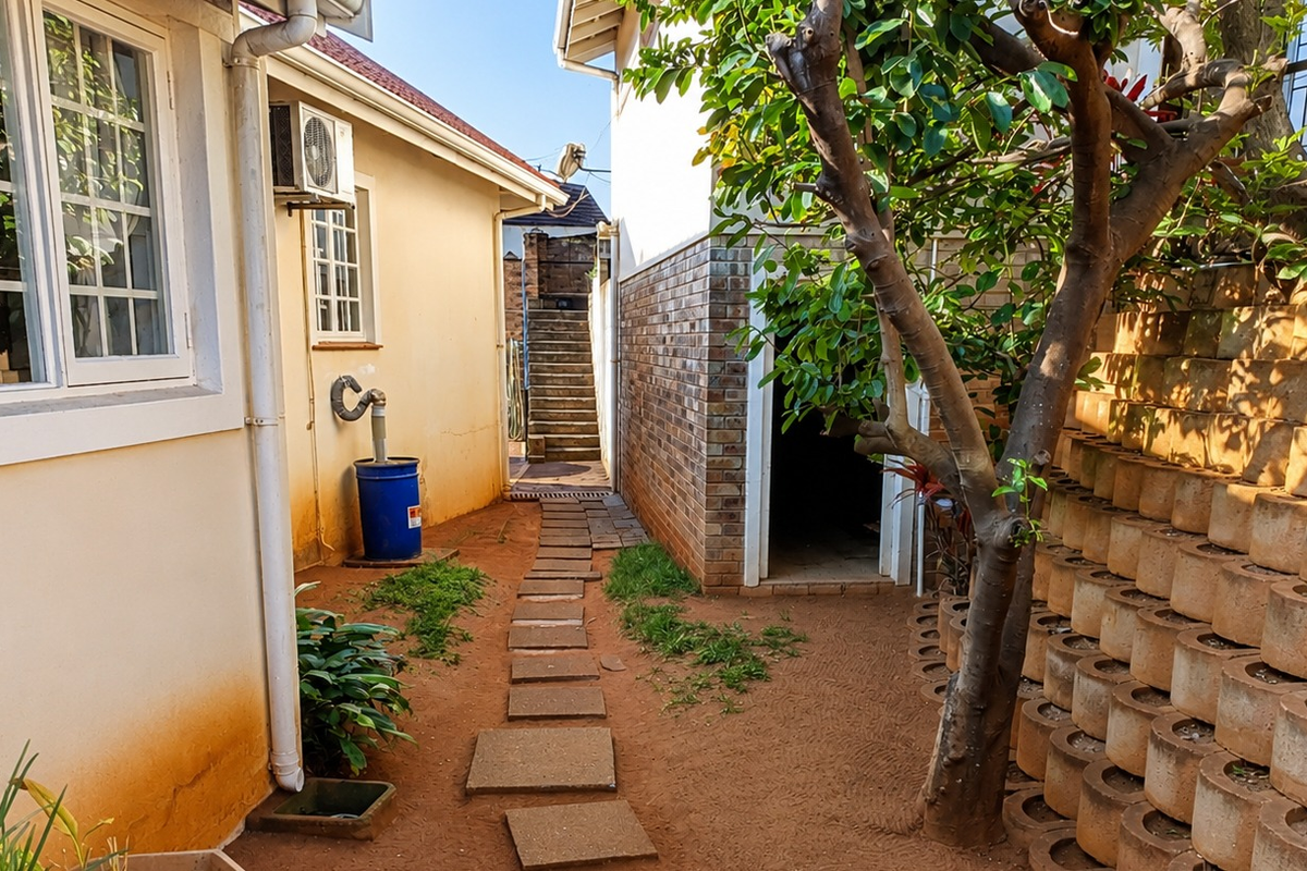 A side exterior pathway featuring a paved walkway, retaining wall, and external building infrastructure including an air conditioning unit and guttering.