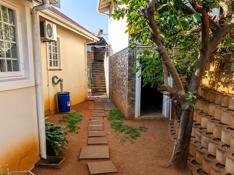 A side exterior pathway featuring a paved walkway, retaining wall, and external building infrastructure including an air conditioning unit and guttering.