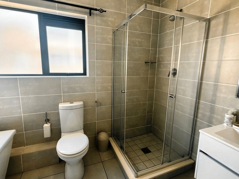 A functional bathroom featuring a glass-enclosed shower, modern vanity with basin, toilet, and neutral floor-to-ceiling tiling.