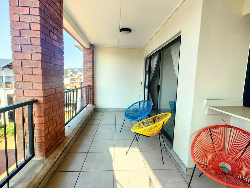 A covered balcony featuring tiled flooring, exposed brick columns, and a metal balustrade, providing an outdoor space accessible via sliding glass doors.