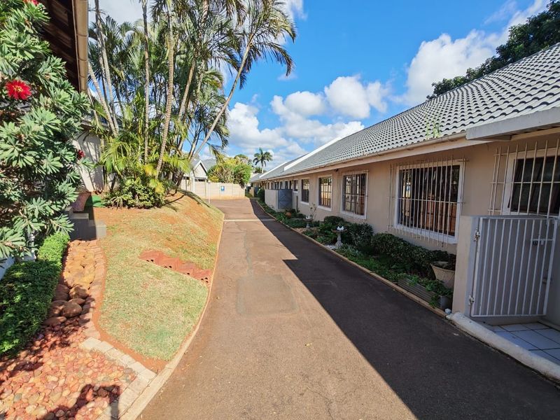 A view of the townhouse complex driveway featuring paved access, established garden landscaping, and residential units with security-fitted windows.