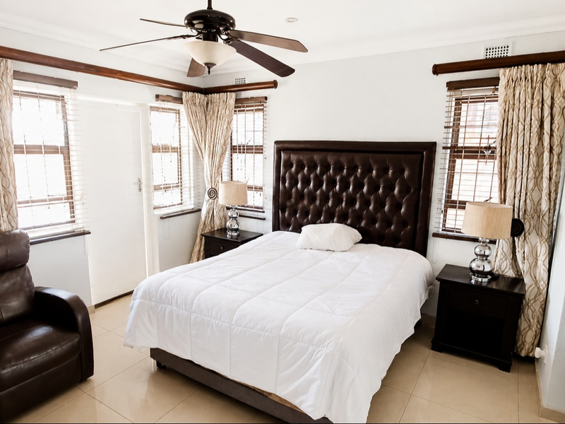 A well-maintained bedroom featuring tiled flooring, a ceiling fan, and large windows with venetian blinds, offering a bright and comfortable living space.