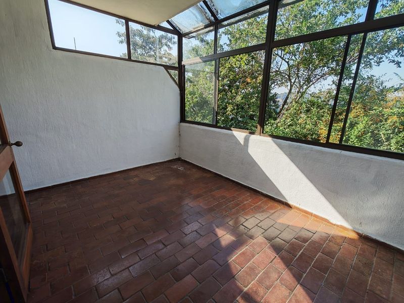 An enclosed sunroom or balcony featuring terracotta tile flooring and large glass windows, offering a bright, versatile space with views of surrounding greenery.
