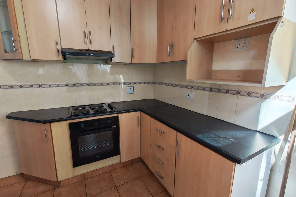 A functional kitchen featuring light wood-tone cabinetry, black countertops, built-in oven, electric hob, and tiled backsplash with ceramic floor tiles.