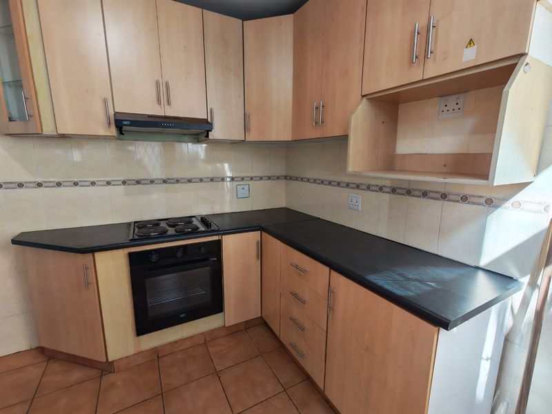 A functional kitchen featuring light wood-tone cabinetry, black countertops, built-in oven, electric hob, and tiled backsplash with ceramic floor tiles.