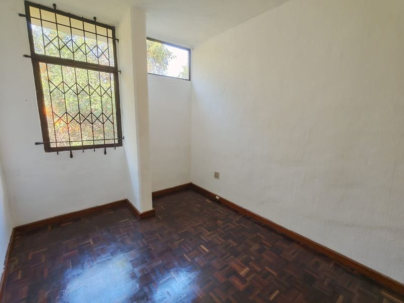 An empty bedroom featuring dark parquet flooring, white textured walls, and a window fitted with security bars.