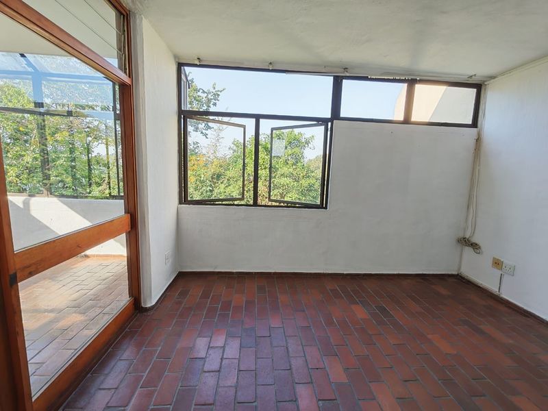 An empty, enclosed balcony featuring classic brick flooring, timber-framed glass doors, and large windows providing natural light and garden views.