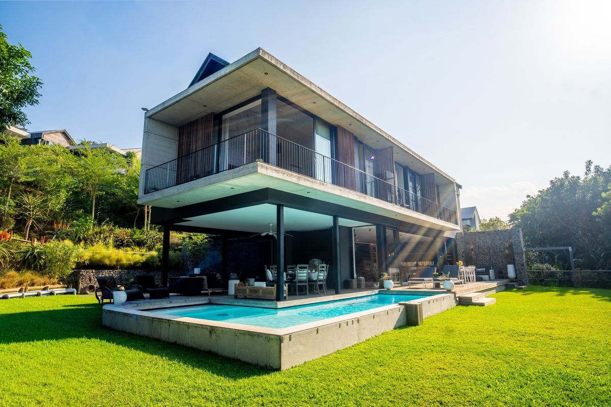 Modern architectural home featuring a concrete structure, elevated balcony, and a sleek swimming pool set within a landscaped garden.