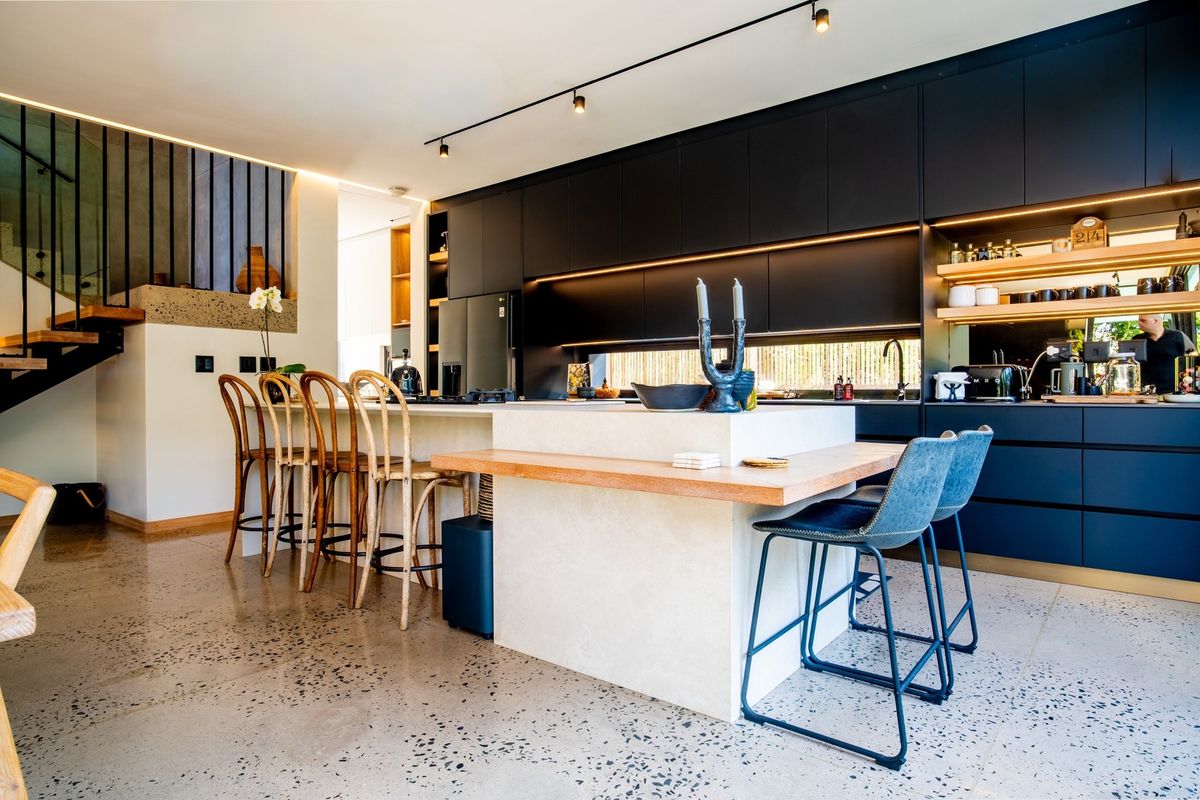 Modern kitchen featuring sleek dark cabinetry, a large stone-topped island, integrated lighting, and polished terrazzo flooring, adjacent to an open staircase.