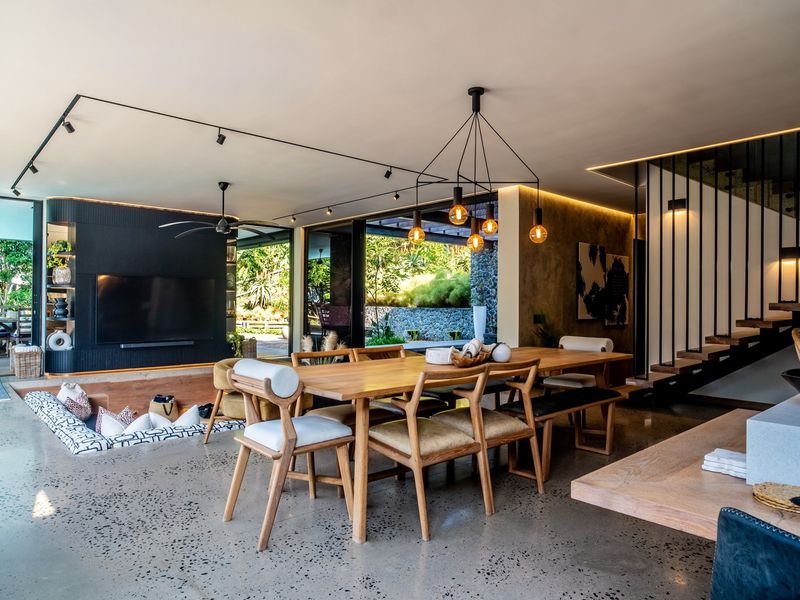 A modern open-plan living and dining area featuring polished concrete floors, a sunken lounge, a striking floating staircase, and seamless indoor-outdoor flow through sliding glass doors.