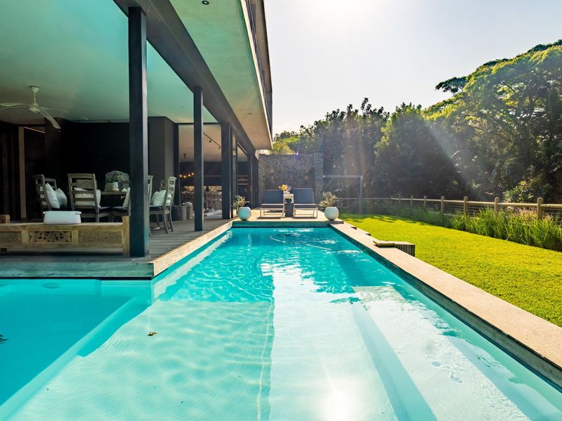 A modern swimming pool area featuring timber decking, a spacious covered patio, and a well-maintained garden with a scenic backdrop.