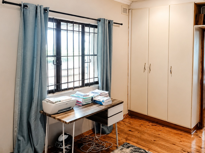 Bedroom featuring polished timber flooring, built-in storage cupboards, and a window with security bars, offering a functional space with potential for cosmetic updates.