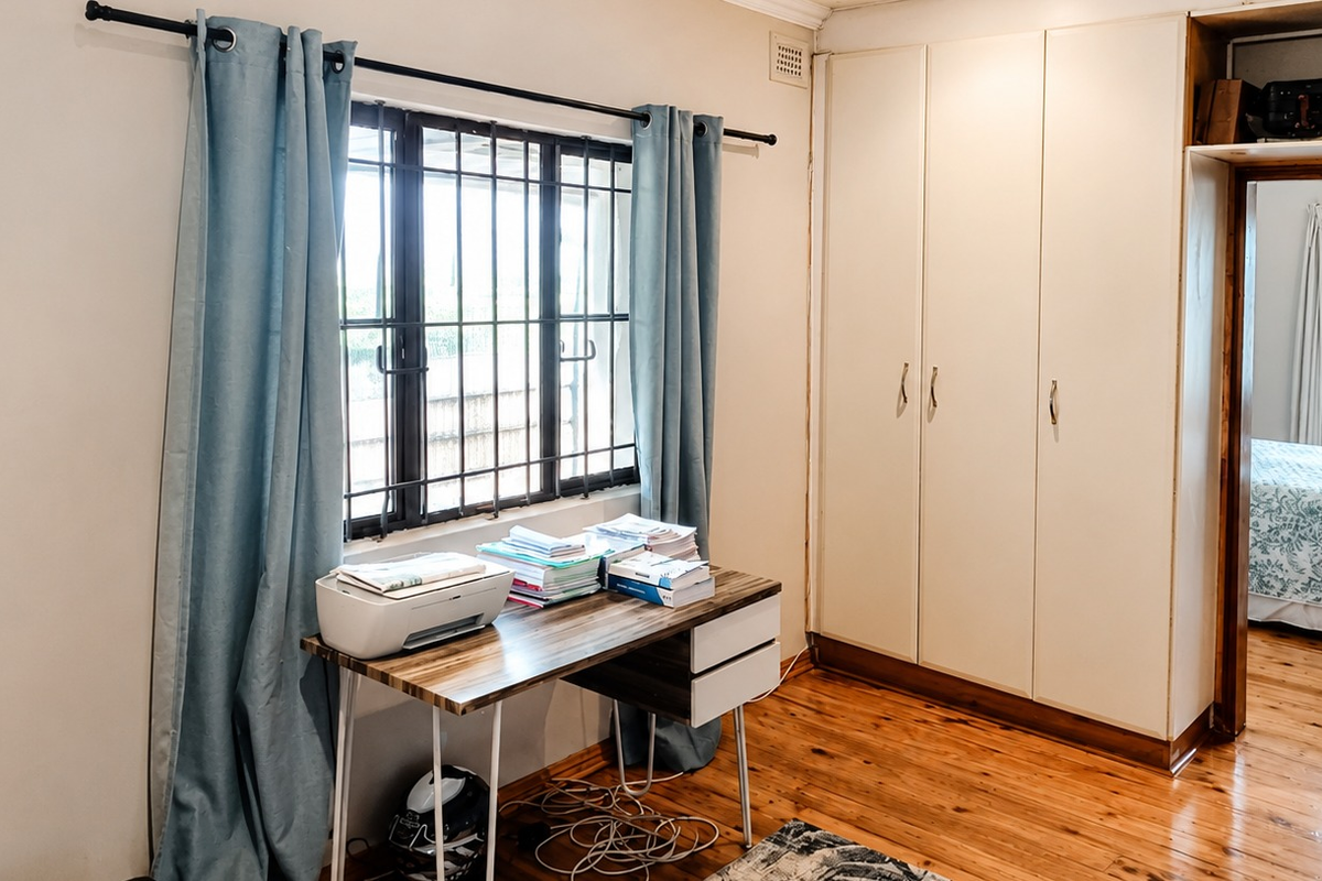Bedroom featuring polished timber flooring, built-in storage cupboards, and a window with security bars, offering a functional space with potential for cosmetic updates.