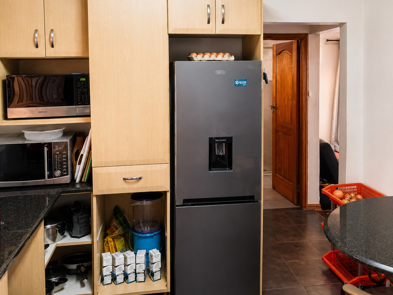 A functional kitchen featuring light wood cabinetry, granite countertops, and a dedicated housing unit for a modern refrigerator, leading into an adjacent room.