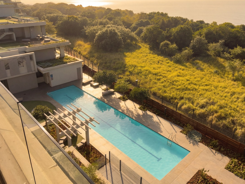 An elevated view of a modern swimming pool area featuring a pergola, paved deck, and glass balustrades, set against a coastal landscape.