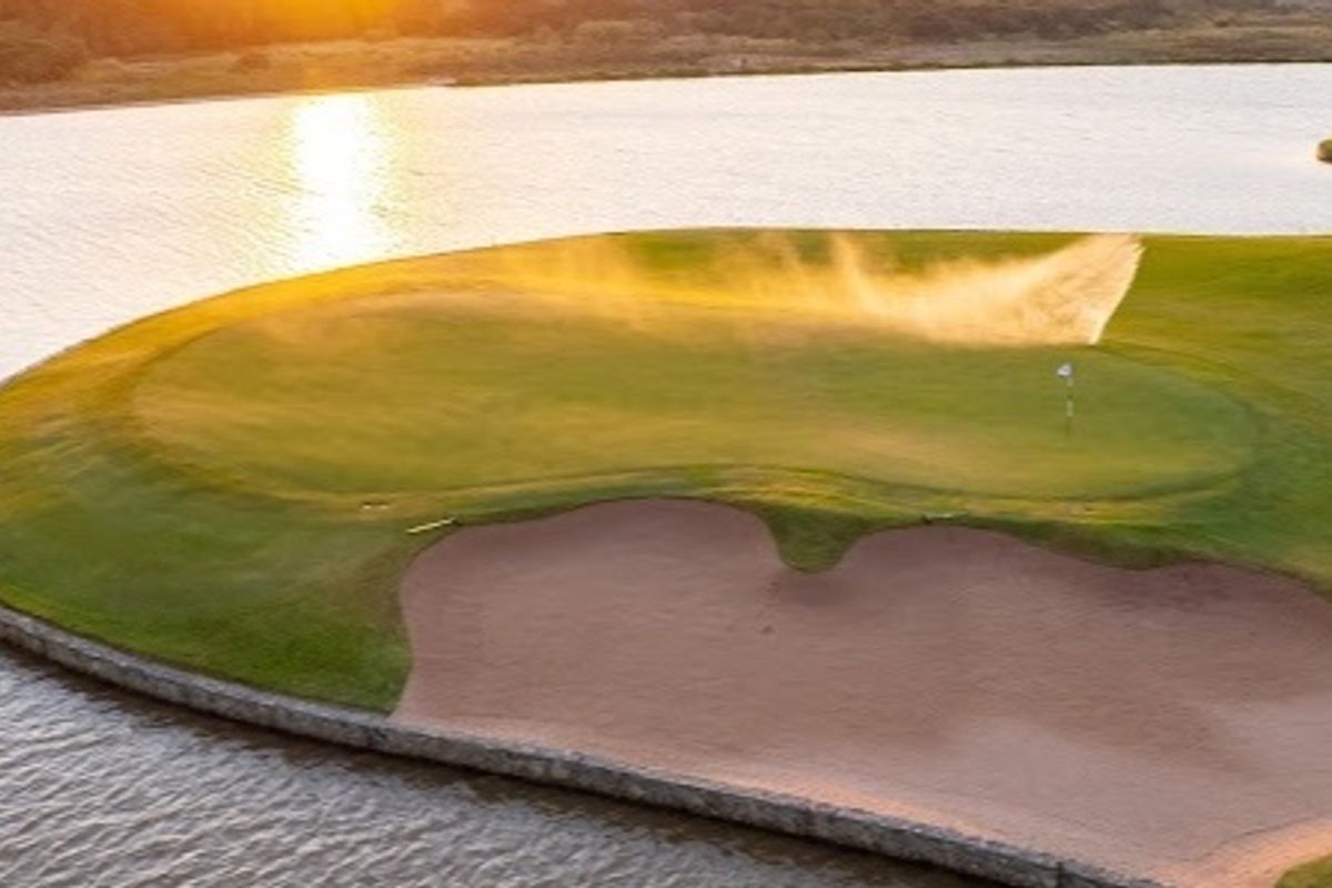 An aerial view of a manicured golf course green situated on a peninsula, featuring a sand bunker and surrounding water hazard at sunset.