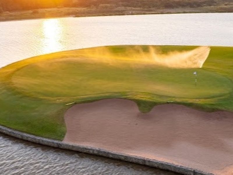 An aerial view of a manicured golf course green situated on a peninsula, featuring a sand bunker and surrounding water hazard at sunset.
