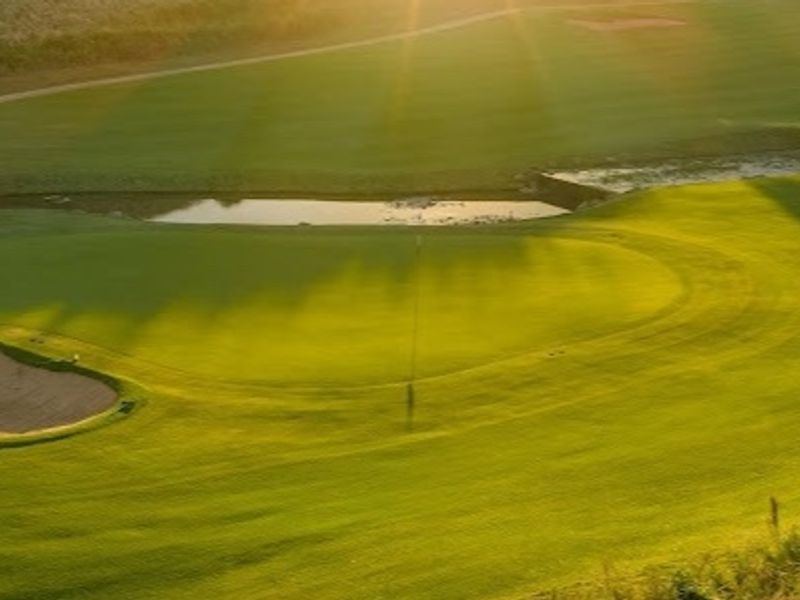 A scenic elevated view of a well-maintained golf course featuring a sand bunker and a water hazard at sunset.