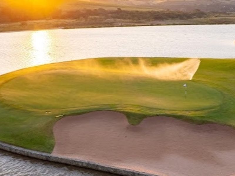 An elevated view of a manicured golf course green featuring a sand bunker and water hazard at sunset.