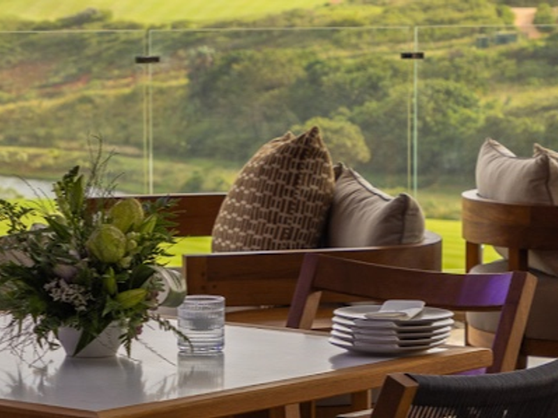 A scenic outdoor patio area featuring a modern glass balustrade overlooking a lush green landscape and water feature.