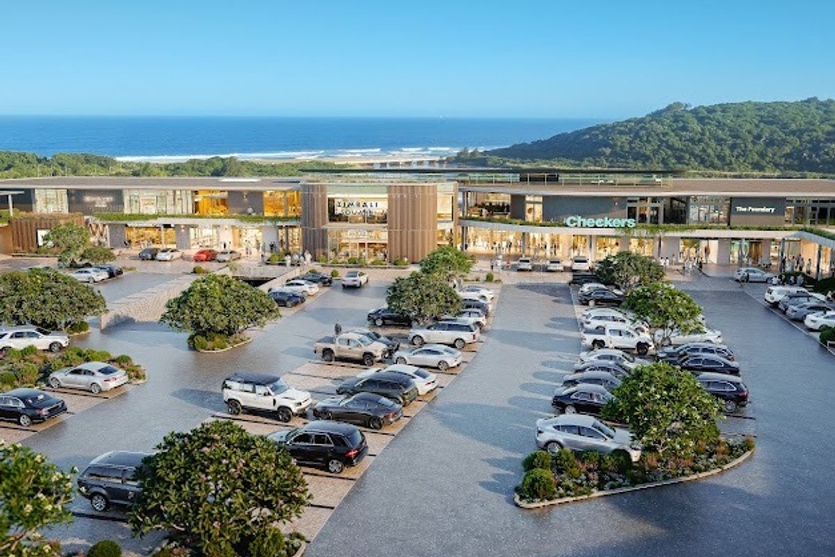 Aerial view of a modern commercial shopping center with extensive paved parking, landscaped greenery, and a scenic coastal backdrop.