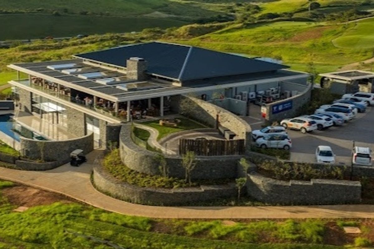 Aerial view of a modern clubhouse facility featuring stone architecture, a swimming pool, and extensive paved parking within a landscaped estate setting.