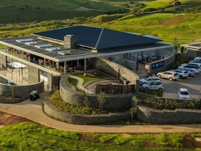 Aerial view of a modern clubhouse facility featuring stone architecture, a swimming pool, and extensive paved parking within a landscaped estate setting.