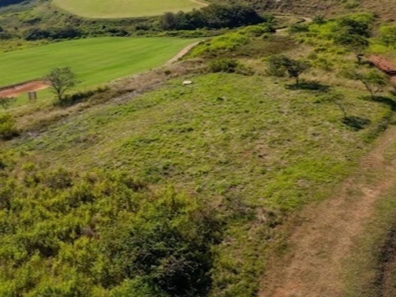 An aerial view of a vacant residential plot featuring sloped terrain, natural grassland, and proximity to a golf course and access road.