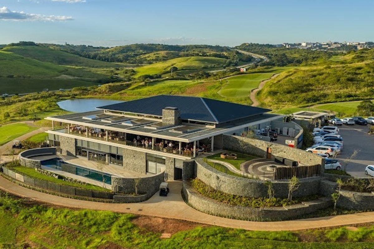 Aerial view of a modern clubhouse facility featuring stone architecture, a swimming pool, and extensive landscaped grounds within a golf estate setting.