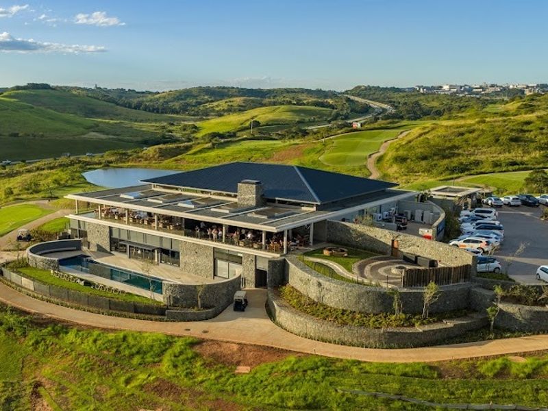 Aerial view of a modern clubhouse facility featuring stone architecture, a swimming pool, and extensive landscaped grounds within a golf estate setting.