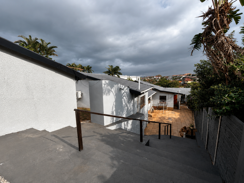 Rear view of the property featuring a tiled patio area, concrete staircase with metal handrail, and surrounding boundary walls under a cloudy sky.