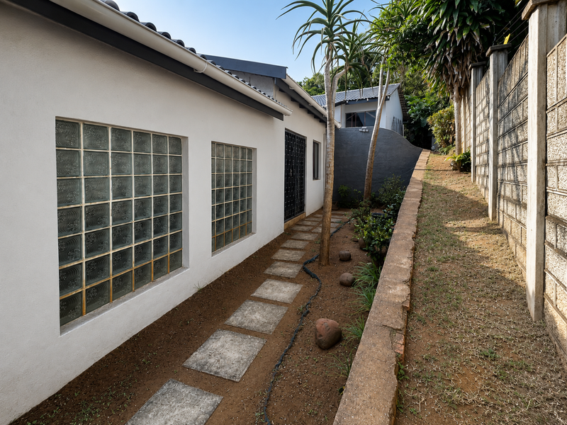 A side exterior view showing a paved walkway, glass block windows, and a retaining wall along the property boundary.