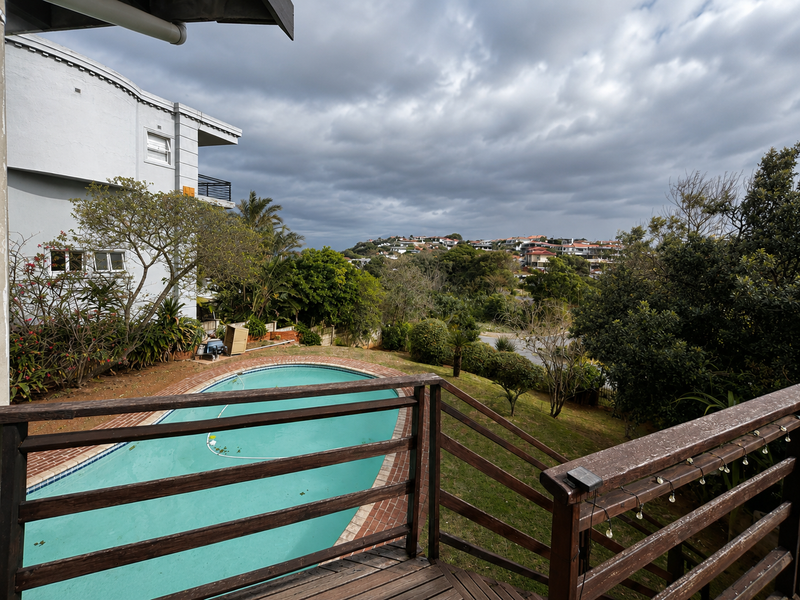 Elevated wooden deck overlooking a swimming pool and surrounding residential landscape with mature trees and cloudy sky.