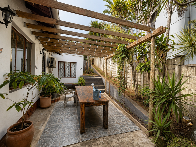 A covered outdoor patio area featuring a wooden pergola with string lighting, tiled flooring, and access to a tiered garden space.