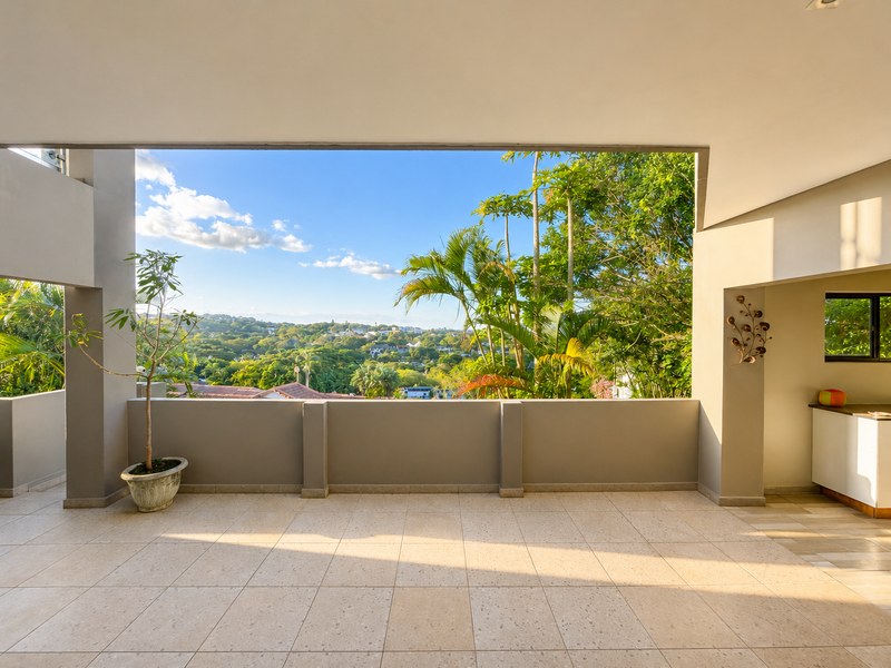 A spacious covered patio featuring tiled flooring and a concrete balustrade, offering expansive elevated views of the surrounding landscape and greenery.