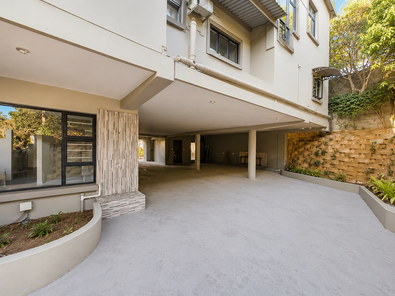 A spacious, covered ground-level parking area featuring a concrete driveway, retaining wall, and modern exterior finishes.