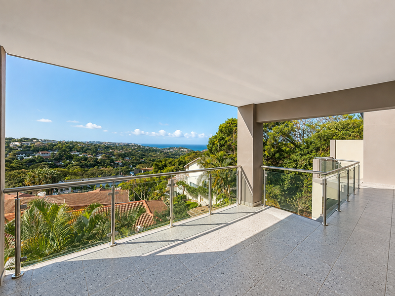 A spacious covered balcony featuring modern glass balustrades, stainless steel railings, and tiled flooring, offering expansive elevated views of the surrounding landscape and ocean.
