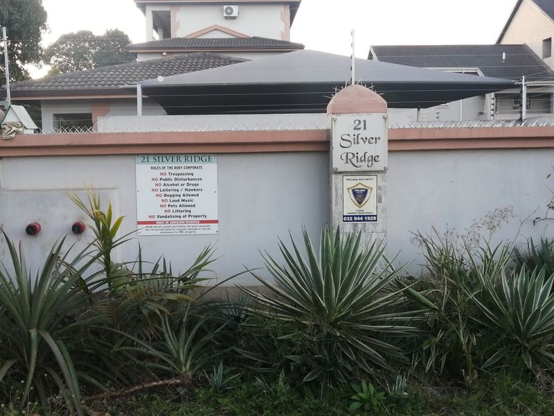 Front view of the apartment complex entrance featuring a secure perimeter wall with electric fencing and a shaded parking area.