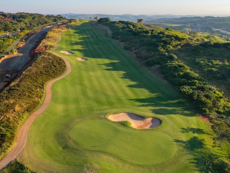 An aerial view of a well-maintained golf course fairway featuring sand bunkers and a paved cart path, set within a scenic landscape.