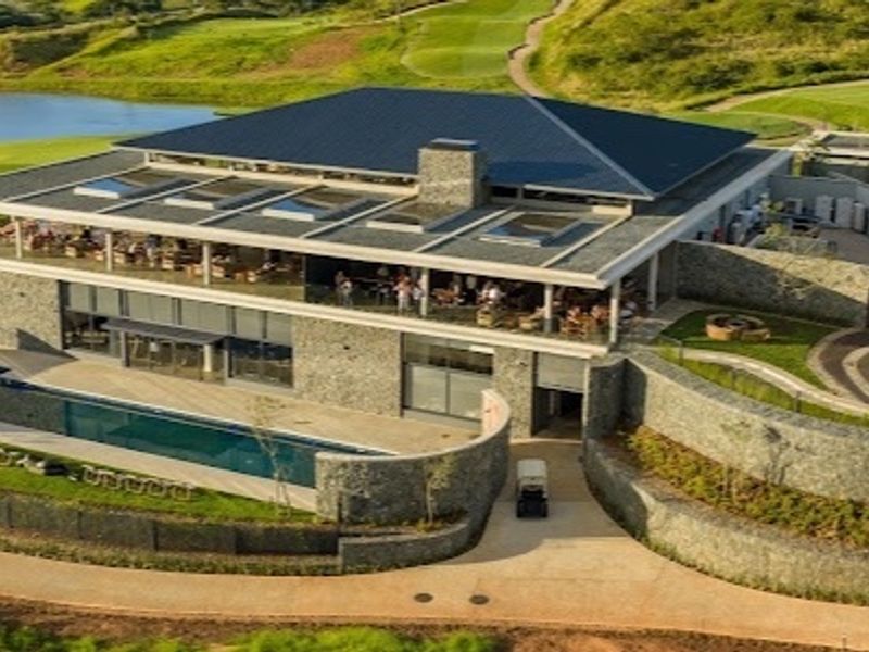 Aerial view of a modern clubhouse facility featuring stone exterior walls, a swimming pool, landscaped grounds, and a large tiled roof with integrated skylights.
