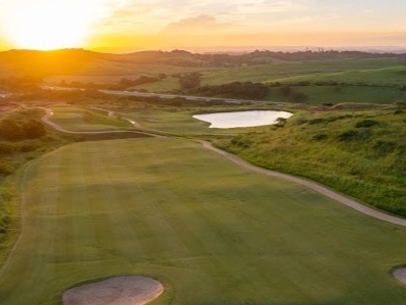 Aerial view of a scenic golf course landscape featuring rolling hills, a water hazard, and sand bunkers at sunset.