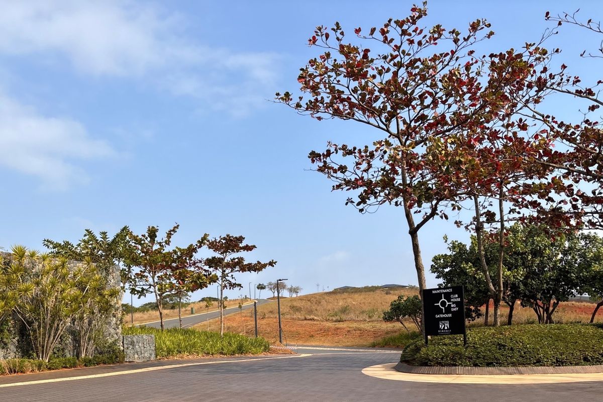 A well-maintained paved entrance road within a residential estate, featuring landscaped gardens, stone retaining walls, and clear directional signage.
