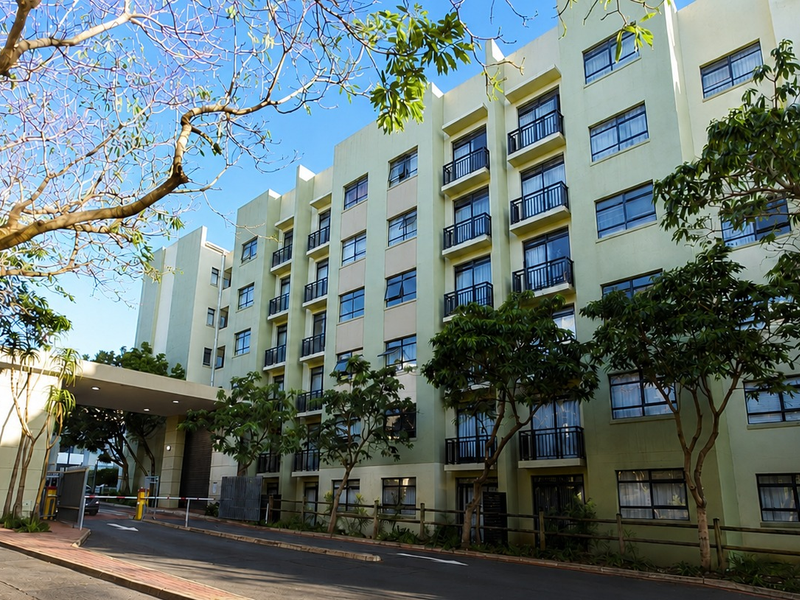 Exterior view of a multi-story apartment complex featuring a paved driveway, covered entrance, and multiple balconies with security railings.