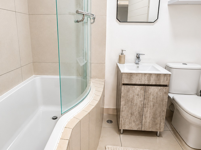 A modern bathroom featuring a curved shower screen over a bathtub, a wood-finish vanity cabinet with a basin, and a standard toilet.