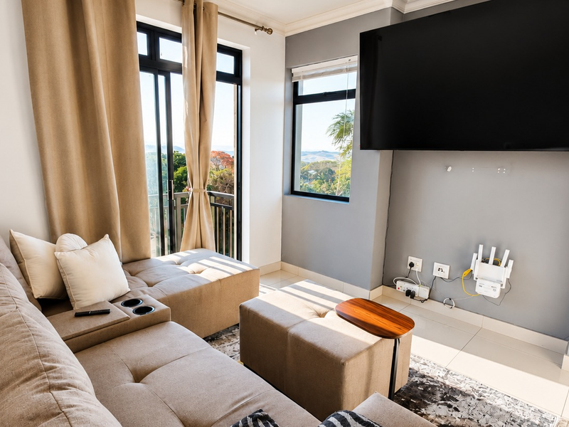 A bright living room featuring tiled floors, large sliding glass doors leading to a balcony, and a wall-mounted television setup.