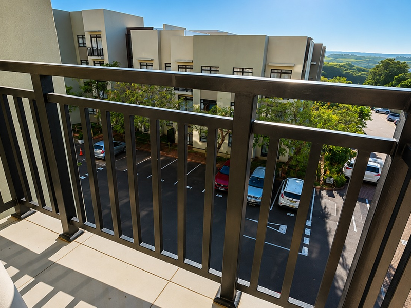 A private balcony featuring a secure metal balustrade and tiled flooring, offering an elevated view over the complex parking area and surrounding landscape.
