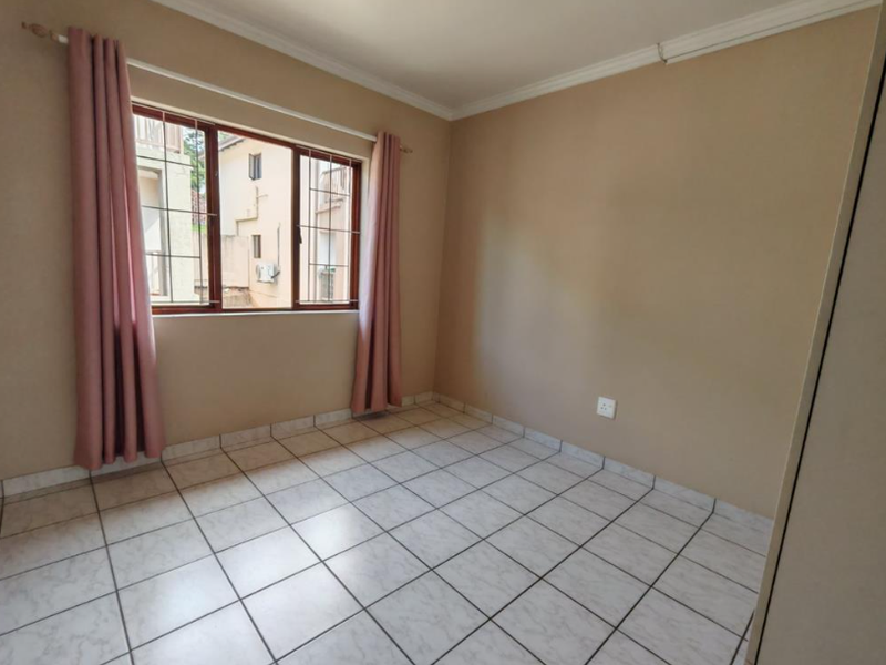 A bright, empty bedroom featuring tiled flooring, built-in cupboards, and security bars on the window.
